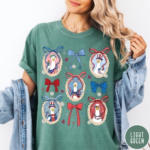 May include: Light green t-shirt featuring a patriotic design with cartoon geese in oval frames. Each goose is dressed in red, white, and blue, with bows and floral accents. The shirt has a relaxed fit and short sleeves.