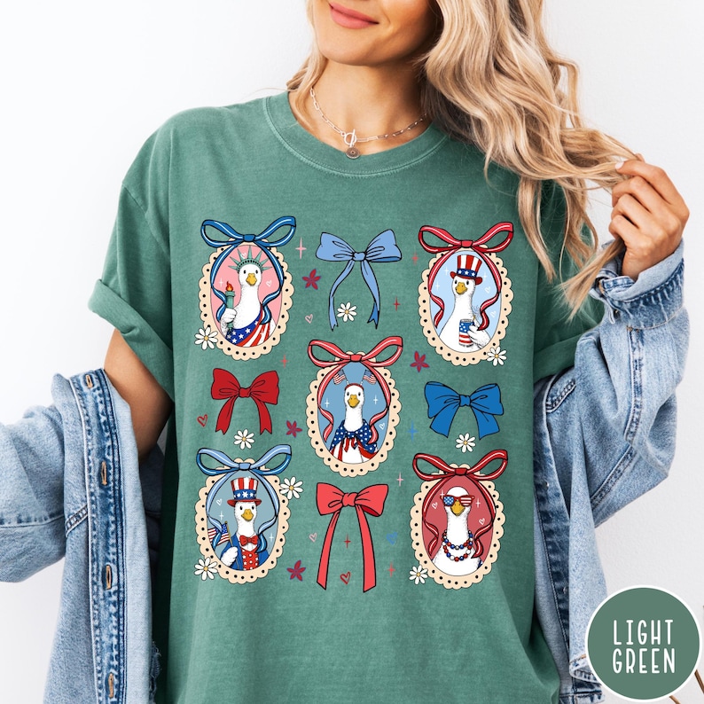 May include: Light green t-shirt featuring a patriotic design with cartoon geese in oval frames. Each goose is dressed in red, white, and blue, with bows and floral accents. The shirt has a relaxed fit and short sleeves.