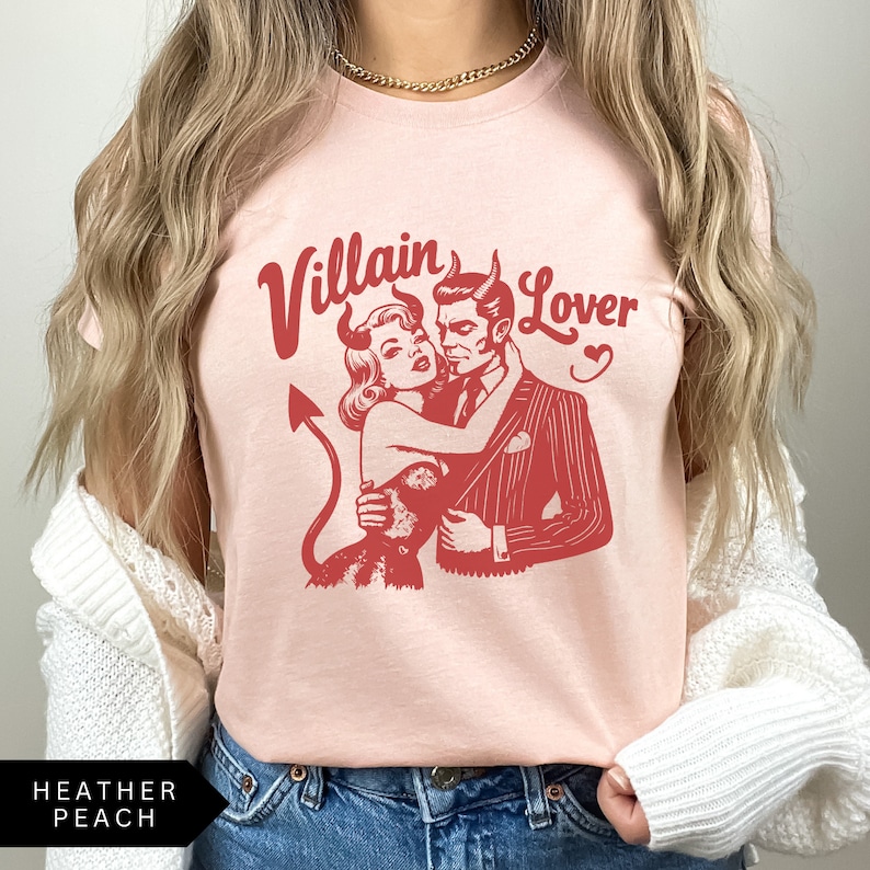 May include: A pink t-shirt with a red graphic of a woman with devil horns and a man in a suit. The text "Villain Lover" is printed in red above the graphic.