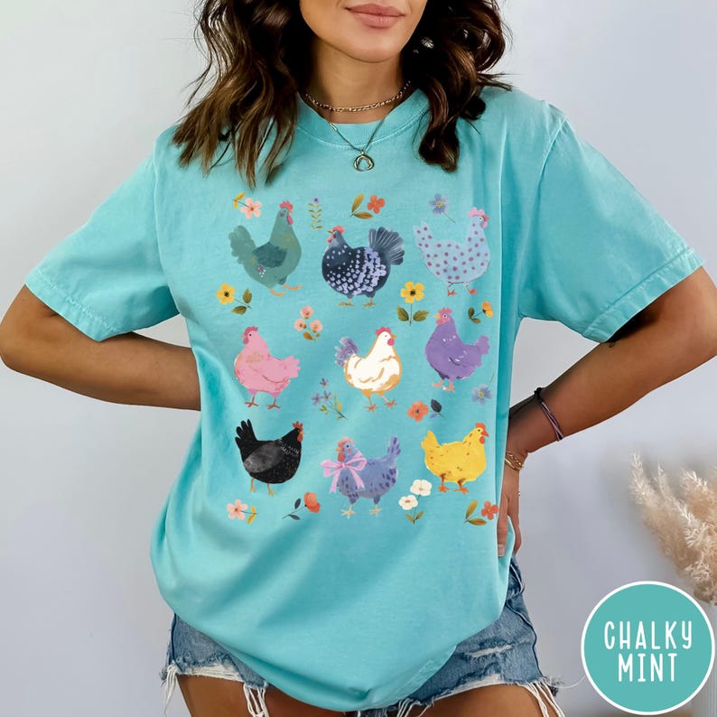 K&ouml;nnte beinhalten: Ein t&uuml;rkisfarbenes T-Shirt mit einem bunten Grafikdruck von H&uuml;hnern. Die H&uuml;hner sind in verschiedenen Posen und Farben dargestellt, darunter rosa, lila, blau und gelb. Das Shirt hat eine lockere Passform und kurze &Auml;rmel.