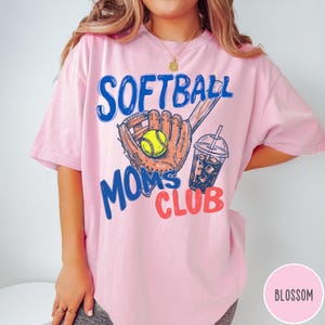 May include: A light pink t-shirt with the words "SOFTBALL MOMS CLUB" in blue and red. The graphic includes a softball glove, a yellow softball, and a drink with ice. The shirt is a comfortable, casual style.