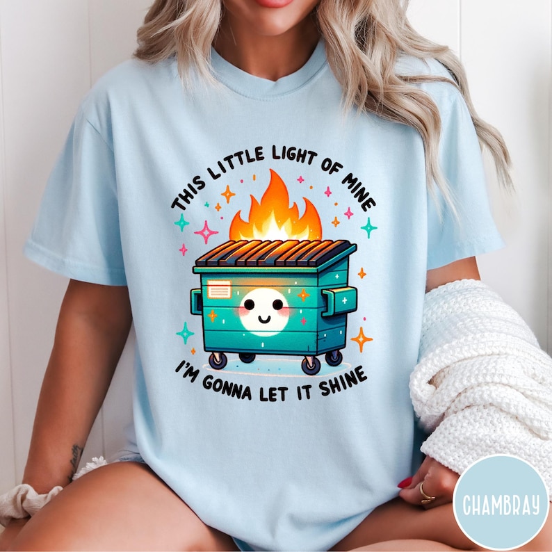May include: A light blue t-shirt with a graphic of a green dumpster with a smiling face and flames coming out of the top. The text "This little light of mine I'm gonna let it shine" is printed on the shirt.