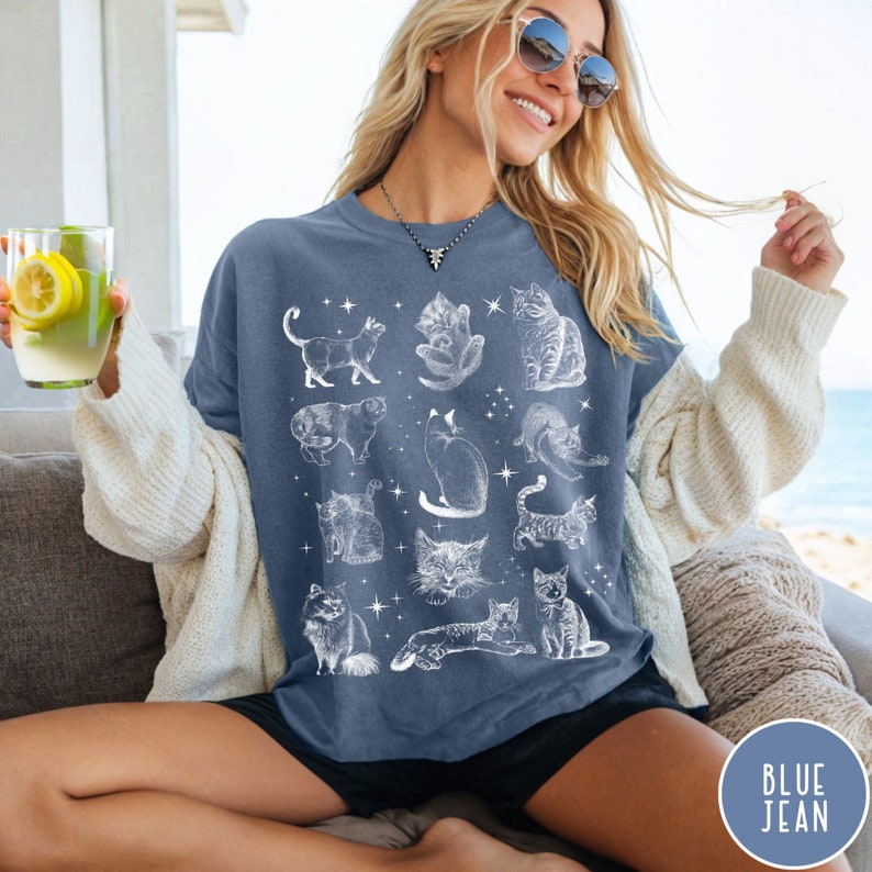 K&ouml;nnte beinhalten: Eine Frau tr&auml;gt ein T-Shirt in Jeansblau mit einem wei&szlig;en Aufdruck von verschiedenen Katzen. Das Shirt hat eine lockere Passform und einen Rundhalsausschnitt.