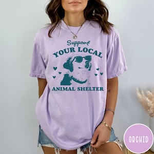 May include: A lavender t-shirt with the text "Support Your Local Animal Shelter" in teal blue lettering. A cartoon dog wearing sunglasses is in the center of the text.  The shirt is worn by a person with a light brown complexion and dark brown hair.