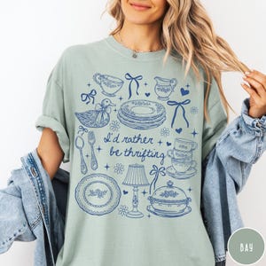 May include: Pale green t-shirt featuring a blue illustration of vintage crockery, including plates, teacups, and a duck. The text "I'd rather be thrifting" is printed in blue. The shirt has a relaxed fit.