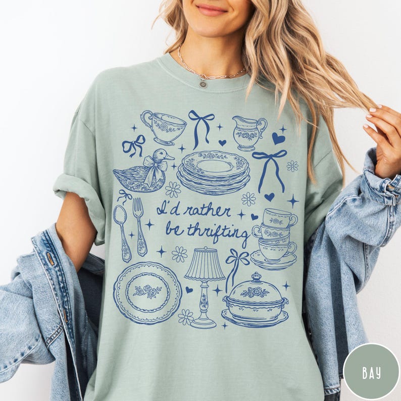 May include: Pale green t-shirt featuring a blue illustration of vintage crockery, including plates, teacups, and a duck. The text "I'd rather be thrifting" is printed in blue. The shirt has a relaxed fit.
