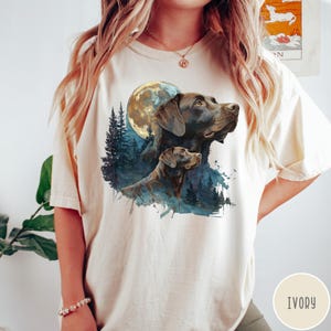 May include: Ivory-colored t-shirt featuring a watercolor illustration of two Labrador Retrievers under a full moon. The design includes a forest silhouette in shades of blue and green. The shirt is a relaxed fit.