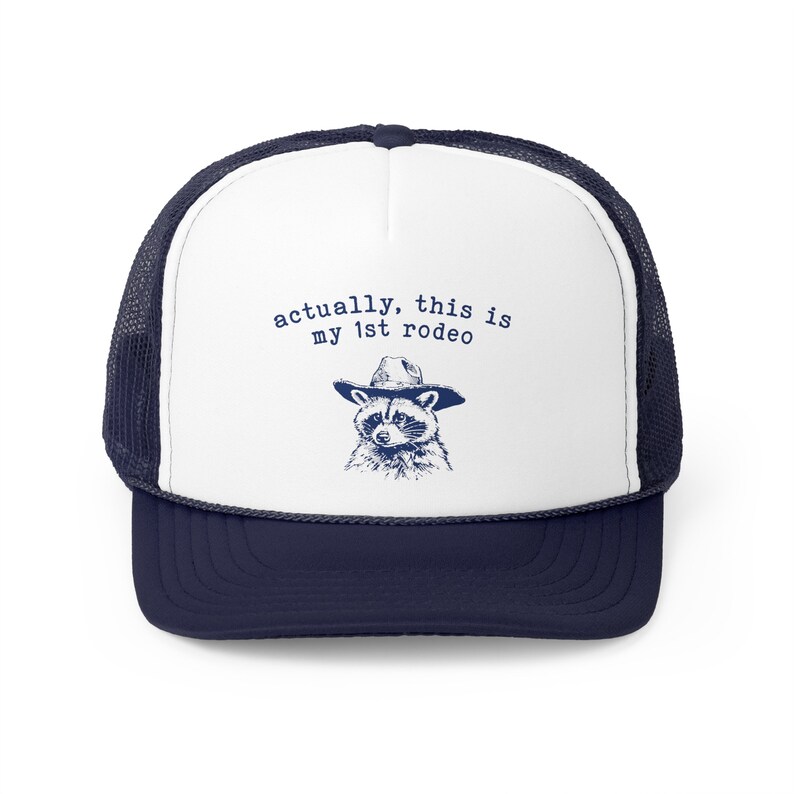 May include: White trucker hat with a navy blue mesh back. The front of the hat features a raccoon wearing a cowboy hat and the text "actually, this is my 1st rodeo".