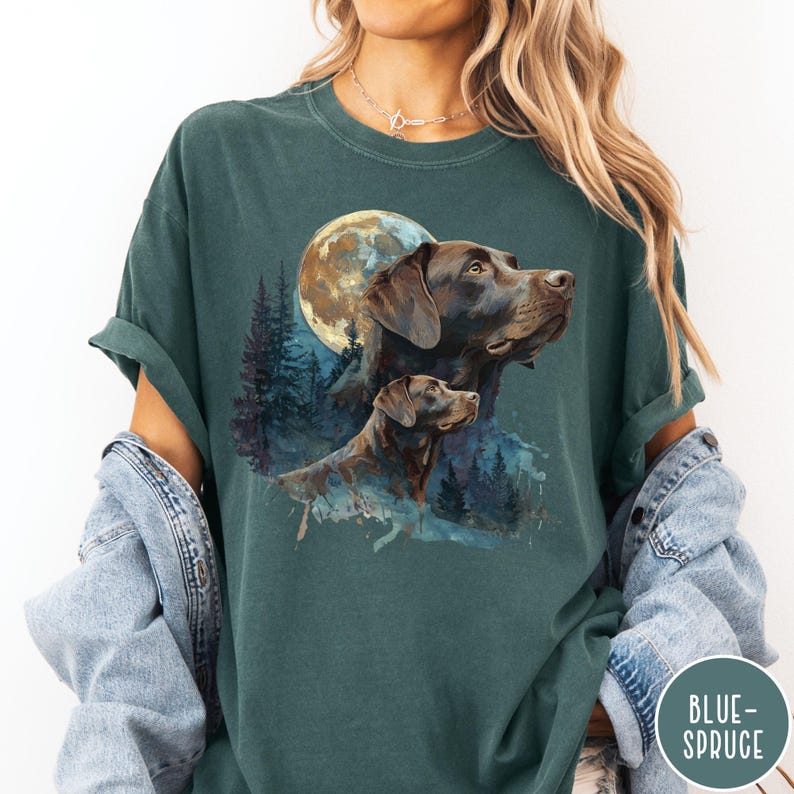 May include: Dark teal t-shirt featuring a watercolor illustration of two Labrador Retrievers with a full moon and forest background. The graphic design includes shades of brown, blue, and white. The shirt has rolled-up sleeves.