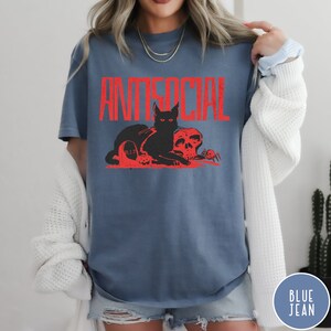 Może przedstawiać: Niebieski T-shirt z napisem "ANTISOCIAL" w czerwonych literach. Czarny kot z czerwonymi oczami siedzi obok czaszki i pająka. Kot znajduje się na nagrobku z napisem "RIP".
