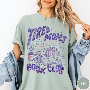 May include: A light green t-shirt with the words "Tired Moms Book Club" in purple script. The design includes illustrations of books, candles, plants, and stars. The text "Running on coffee, chaos & chapters" is also included.