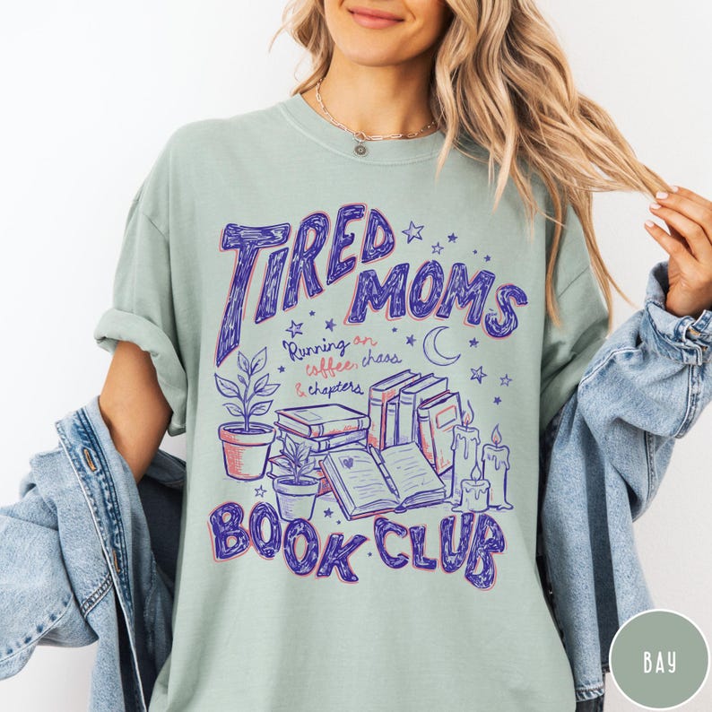 May include: A light green t-shirt with the words "Tired Moms Book Club" in purple script. The design includes illustrations of books, candles, plants, and stars. The text "Running on coffee, chaos & chapters" is also included.