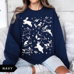 May include: Navy blue sweatshirt with a repeating pattern of white rabbits, stars, and planets. The rabbits are in different poses, creating a whimsical design. The sweatshirt is a comfortable, casual garment, perfect for everyday wear.