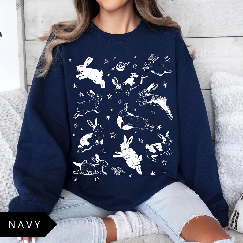 May include: Navy blue sweatshirt with a repeating pattern of white rabbits, stars, and planets. The rabbits are in different poses, creating a whimsical design. The sweatshirt is a comfortable, casual garment, perfect for everyday wear.