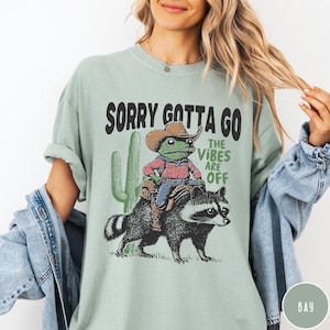 May include: Sage green t-shirt with a graphic featuring a frog in a cowboy hat riding a raccoon, with the text "SORRY GOTTA GO THE VIBES ARE OFF". A cactus is also featured in the graphic.