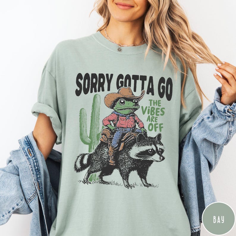 May include: Sage green t-shirt with a graphic featuring a frog in a cowboy hat riding a raccoon, with the text "SORRY GOTTA GO THE VIBES ARE OFF". A cactus is also featured in the graphic.