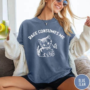 Puede incluir: Una camiseta azul jean con un gr&aacute;fico blanco de un gatito con un lazo y una mariposa. El texto "Rage Consumes Me" est&aacute; impreso en blanco en la camiseta.