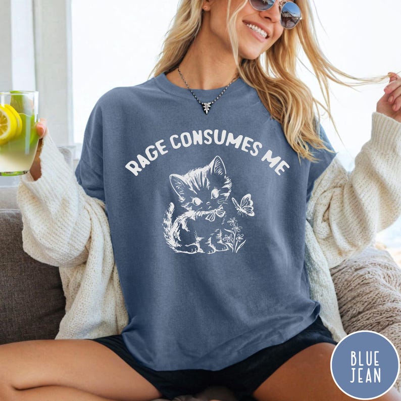Puede incluir: Una camiseta azul jean con un gr&aacute;fico blanco de un gatito con un lazo y una mariposa. El texto "Rage Consumes Me" est&aacute; impreso en blanco en la camiseta.
