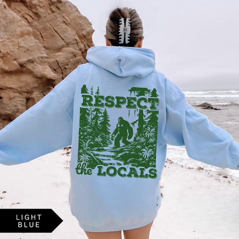 May include: Light blue hooded sweatshirt with a green graphic print of a Bigfoot-like creature in a forest setting. The graphic includes the text "Respect the Locals".