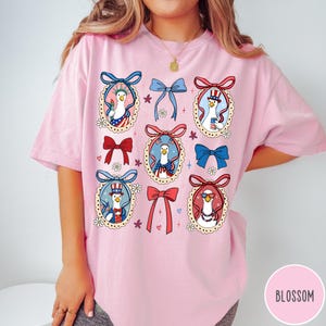 May include: Pink t-shirt featuring a whimsical design of geese in patriotic attire, framed by decorative bows and floral accents. The geese are depicted with sunglasses, top hats, and holding the Statue of Liberty. The word "BLOSSOM" is printed on the bottom right.
