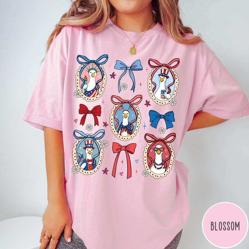 May include: Pink t-shirt featuring a whimsical design of geese in patriotic attire, framed by decorative bows and floral accents. The geese are depicted with sunglasses, top hats, and holding the Statue of Liberty. The word "BLOSSOM" is printed on the bottom right.