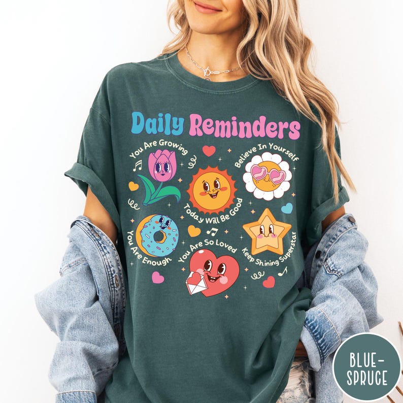 May include: Dark teal t-shirt with the words "Daily Reminders" in a retro font. The shirt features colorful illustrations of a flower, sun, donut, star, and heart, each with positive affirmations. The shirt is a comfortable, casual style.