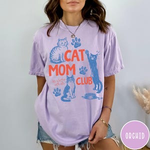 K&ouml;nnte beinhalten: Lilafarbenes T-Shirt mit einem Grafikdesign. Das Design zeigt die Worte "CAT MOM CLUB" in Koralle und Blau, zusammen mit Illustrationen von Katzen, Pfotenabdr&uuml;cken und dem Satz "life is better with cats."