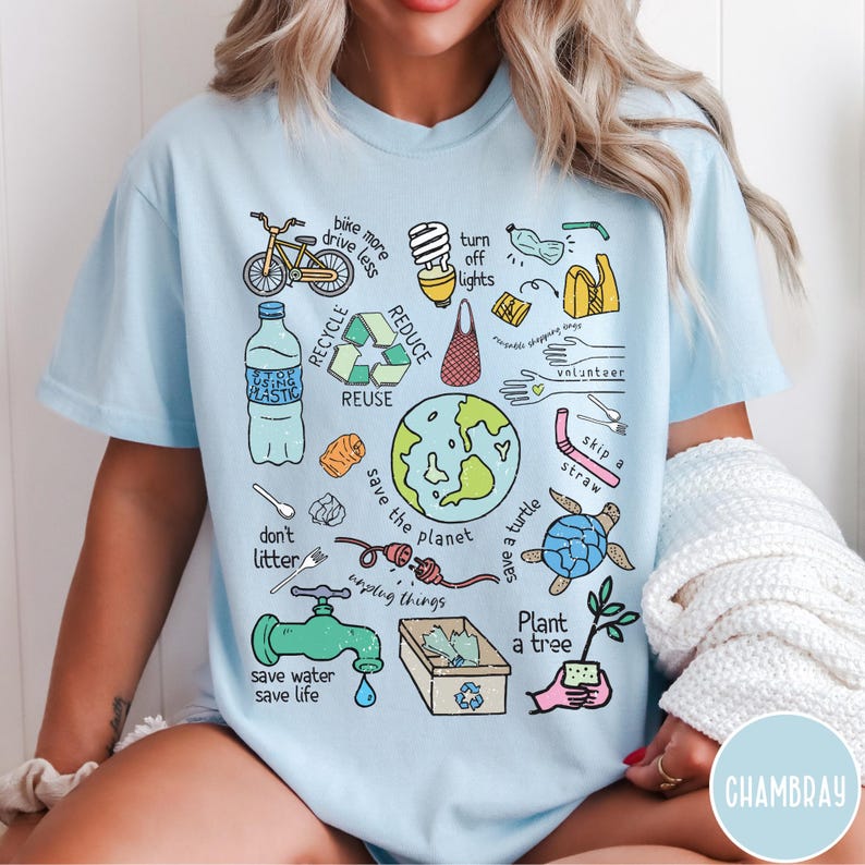 May include: Light blue t-shirt featuring a graphic print of various illustrations related to environmentalism. The illustrations include a bicycle, a lightbulb, a reusable bag, a water bottle, a recycling symbol, a globe, a turtle, a faucet, a tree, and a straw. The text on the shirt includes "bike more drive less", "turn off lights", "reduce", "recycle", "reuse", "save the planet", "don't litter", "save water save life", "skip a straw", "save a turtle", "plant a tree", and "unplug things".