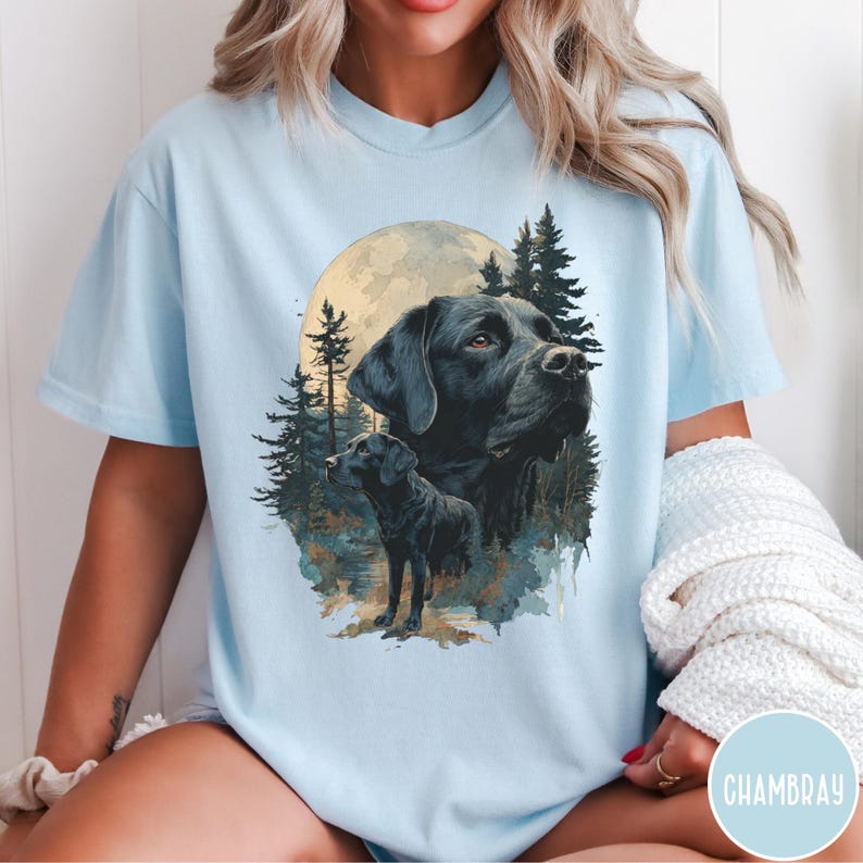 May include: Light blue t-shirt featuring a black Labrador Retriever illustration. The design includes a large full moon, pine trees, and a smaller dog silhouette. The shirt has short sleeves and a relaxed fit.