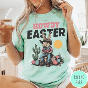Puede incluir: Una camiseta verde claro con las palabras "HOWDY EASTER" en rosa y negro. El gr&aacute;fico presenta un conejito con sombrero de vaquero sentado en una roca con huevos de Pascua y un cactus. Tambi&eacute;n hay un c&iacute;rculo amarillo.