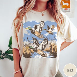 May include: Ivory t-shirt with a retro design of ducks flying over a marsh. The illustration shows ducks with green heads and brown bodies, with wings spread, set against a blue sky and reeds. The word 'IVORY' is printed on the bottom right.