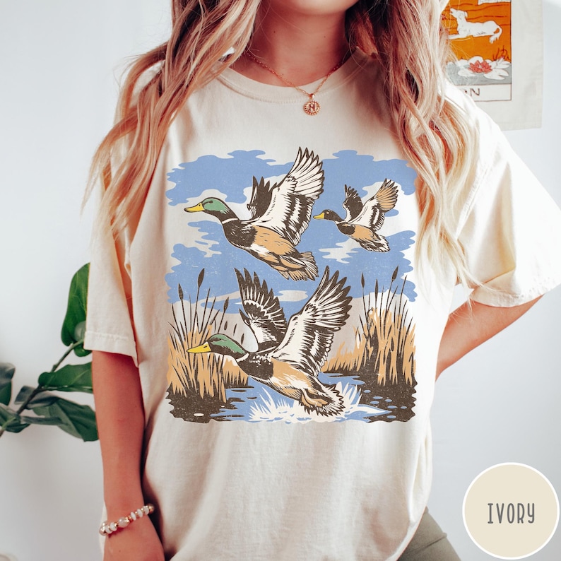 May include: Ivory t-shirt with a retro design of ducks flying over a marsh. The illustration shows ducks with green heads and brown bodies, with wings spread, set against a blue sky and reeds. The word 'IVORY' is printed on the bottom right.