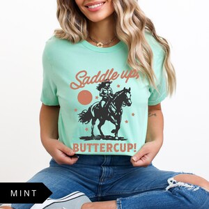 May include: A mint green t-shirt with a graphic of a woman riding a horse. The text "Saddle up Buttercup!" is printed on the shirt.