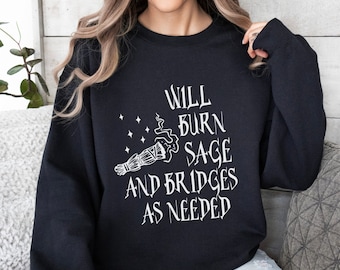 Puede incluir: Sudadera negra con texto blanco "Will Burn Sage and Bridges as Needed". El diseño incluye un manojo de salvia y estrellas. La sudadera es de cuello redondo.