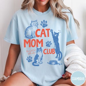 K&ouml;nnte beinhalten: Hellblaues T-Shirt mit einem Grafikdesign. Das Design enth&auml;lt den Text "CAT MOM CLUB" in Orange, Katzenillustrationen und Pfotenabdr&uuml;cke. Der Satz "life is better with cats" ist ebenfalls enthalten.