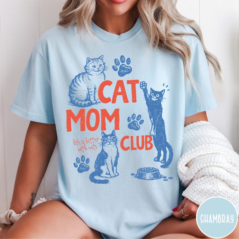 K&ouml;nnte beinhalten: Hellblaues T-Shirt mit einem Grafikdesign. Das Design enth&auml;lt den Text "CAT MOM CLUB" in Orange, Katzenillustrationen und Pfotenabdr&uuml;cke. Der Satz "life is better with cats" ist ebenfalls enthalten.
