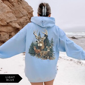 Op de afbeelding: Lichtblauwe hoodie met een hertenafbeelding op de rug. De afbeelding toont een hert met groot gewei, twee kleinere herten en dennenbomen. De tekst "LIGHT BLUE" staat in een zwarte pijl-vormige doos onderaan.