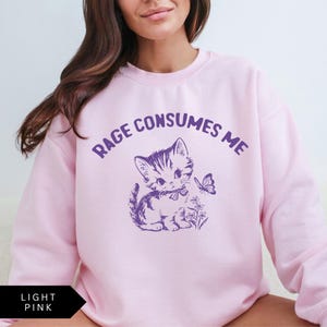 以下が含まれることがあります： 薄いピンクのトレーナーに、紫色の文字で「RAGE CONSUMES ME」と書かれています。紫色のリボンをつけた漫画の猫が、目の前の蝶を見ています。