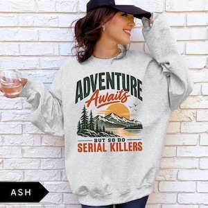 May include: An ash gray crewneck sweatshirt featuring a graphic print of a mountain landscape with a lake, pine trees, and a sunset. The text reads ADVENTURE Awaits BUT SO DO SERIAL KILLERS in dark green and orange lettering across the chest area.