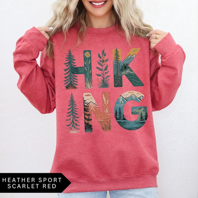 Puede incluir: Una sudadera roja con la palabra "HIKING" en un dise&ntilde;o con tem&aacute;tica de la naturaleza. Las letras est&aacute;n rellenas de im&aacute;genes de &aacute;rboles, monta&ntilde;as y paisajes. La esquina inferior izquierda de la imagen tiene el texto "HEATHER SPORT SCARLET RED".