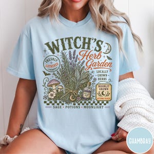 May include: A light blue t-shirt featuring a graphic print with the text "Witch's Herb Garden," "Freshly Gathered," "Locally Grown Herbs," "Moonlit Grown," "Autumn Picked," and "Sage, Potions, Moonlight." The design includes herbs, mushrooms, and a bottle.