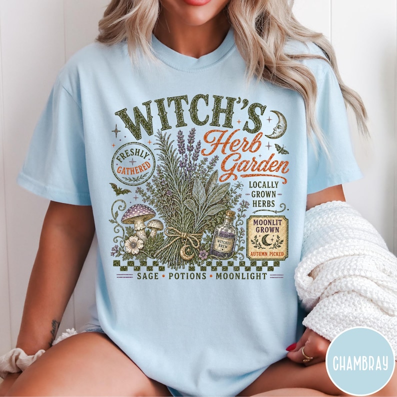 May include: A light blue t-shirt featuring a graphic print with the text "Witch's Herb Garden," "Freshly Gathered," "Locally Grown Herbs," "Moonlit Grown," "Autumn Picked," and "Sage, Potions, Moonlight." The design includes herbs, mushrooms, and a bottle.
