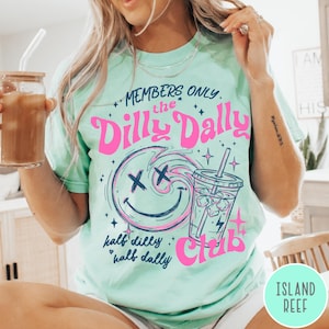May include: A light green t-shirt with the text "MEMBERS ONLY the Dilly Dally Club" in pink and blue. The design includes a smiley face with an "X" for eyes and a drink with ice cubes. The words "half dilly, half dally" are also printed on the shirt.