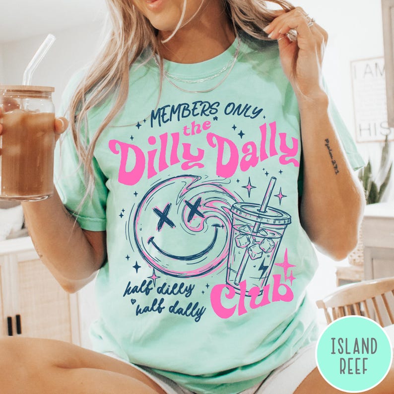 May include: A light green t-shirt with the text "MEMBERS ONLY the Dilly Dally Club" in pink and blue. The design includes a smiley face with an "X" for eyes and a drink with ice cubes. The words "half dilly, half dally" are also printed on the shirt.