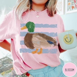 May include: A light pink t-shirt featuring a graphic of a mallard duck with a green head, brown body, and orange feet, set against horizontal blue distressed stripes. The shirt is paired with light blue pants and a white mug with a yellow smiley face.