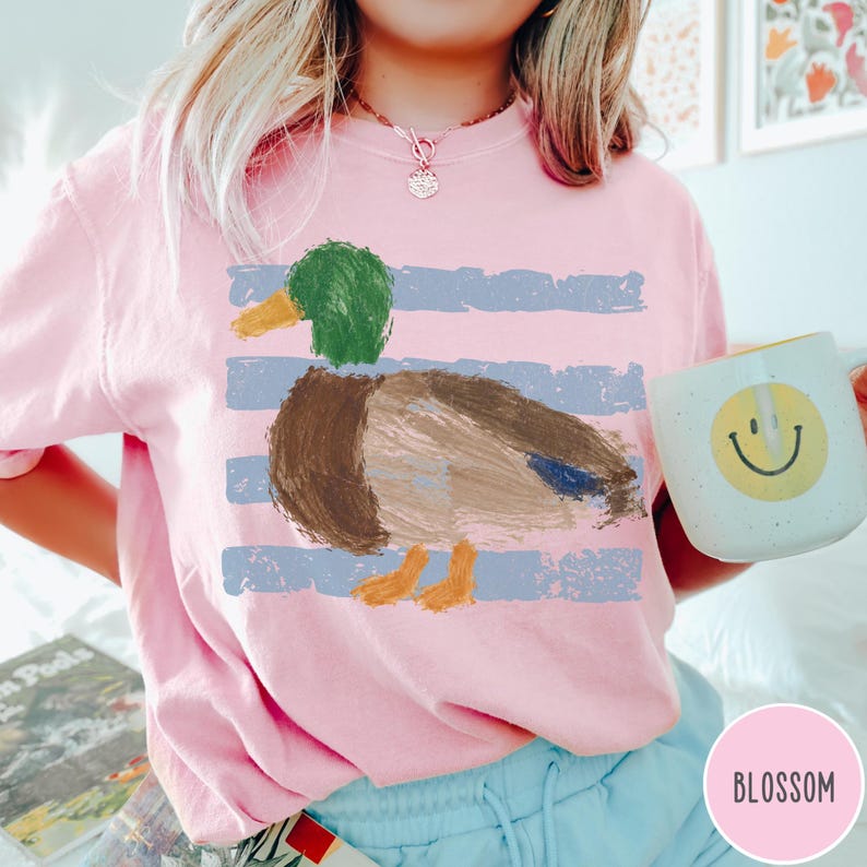 May include: A light pink t-shirt featuring a graphic of a mallard duck with a green head, brown body, and orange feet, set against horizontal blue distressed stripes. The shirt is paired with light blue pants and a white mug with a yellow smiley face.