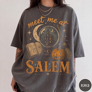 May include: A charcoal gray short-sleeve t-shirt featuring a graphic print with the text meet me at SALEM. The design includes a crystal ball with a house inside, an open book titled SPELLS & CHARMS, a crescent moon, a pumpkin, and a broomstick.
