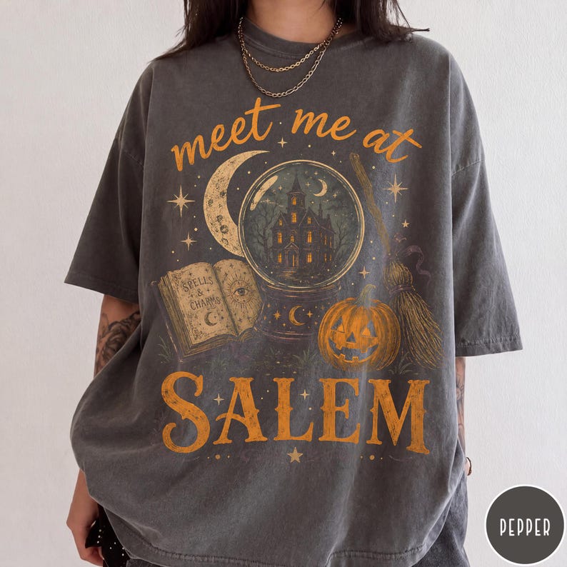 May include: A charcoal gray short-sleeve t-shirt featuring a graphic print with the text meet me at SALEM. The design includes a crystal ball with a house inside, an open book titled SPELLS & CHARMS, a crescent moon, a pumpkin, and a broomstick.