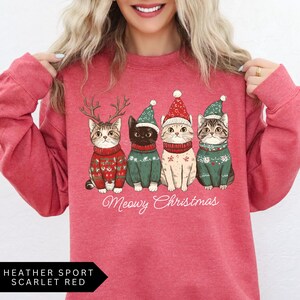 May include: A heather sport scarlet red sweatshirt featuring four cartoon cats in Christmas sweaters and hats. The cats are wearing reindeer antlers, a green hat, a red hat, and a green hat. The text "Meowy Christmas" is below the cats.