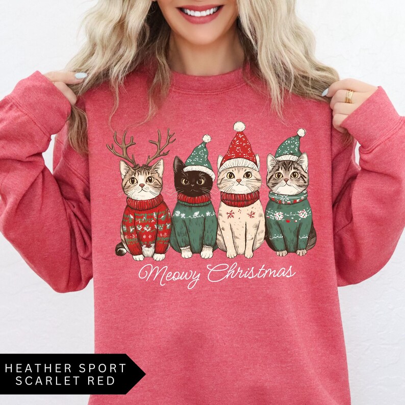 May include: A heather sport scarlet red sweatshirt featuring four cartoon cats in Christmas sweaters and hats. The cats are wearing reindeer antlers, a green hat, a red hat, and a green hat. The text "Meowy Christmas" is below the cats.